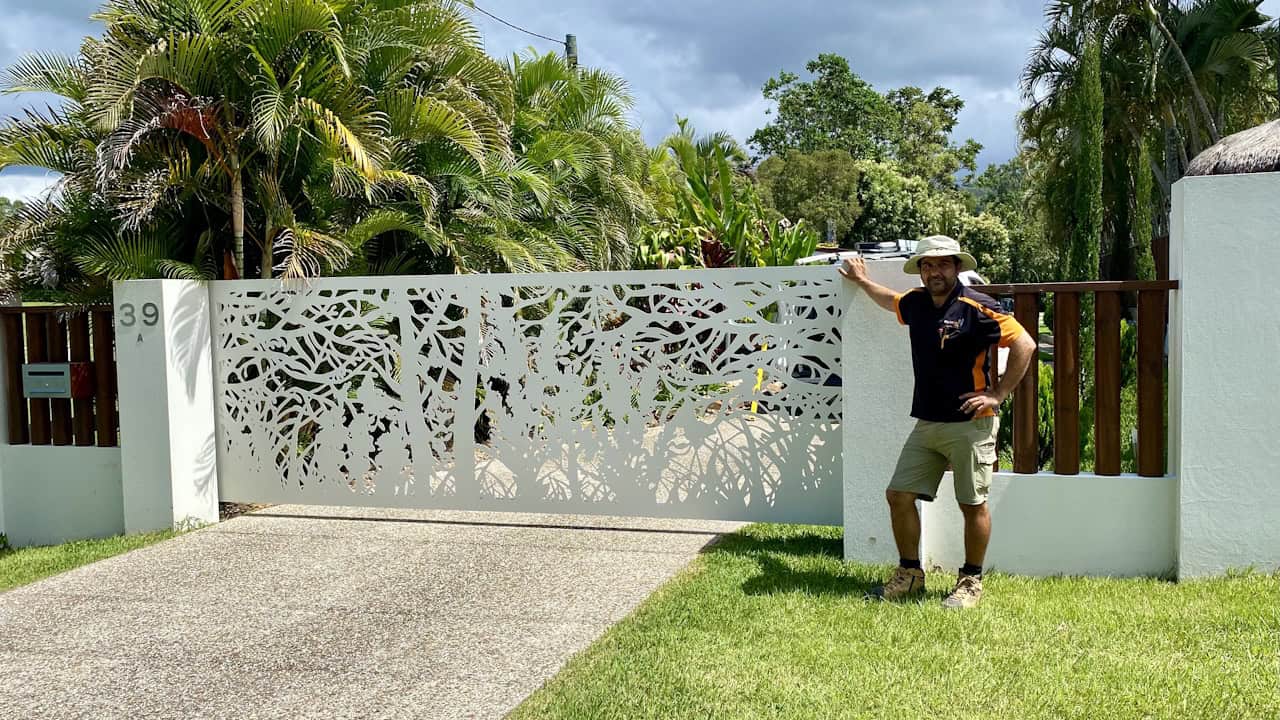 A man in a hat and black shirt poses beside a decorative white metal gate with an intricate leaf cutout pattern, set between concrete pillars with tropical plants behind.