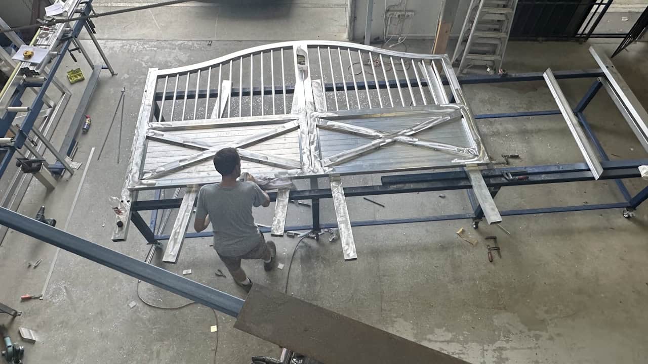 A person kneels in a workshop, working on a large white wooden gate with decorative X-pattern panels and vertical slats, surrounded by metal frames and tools on a concrete floor.