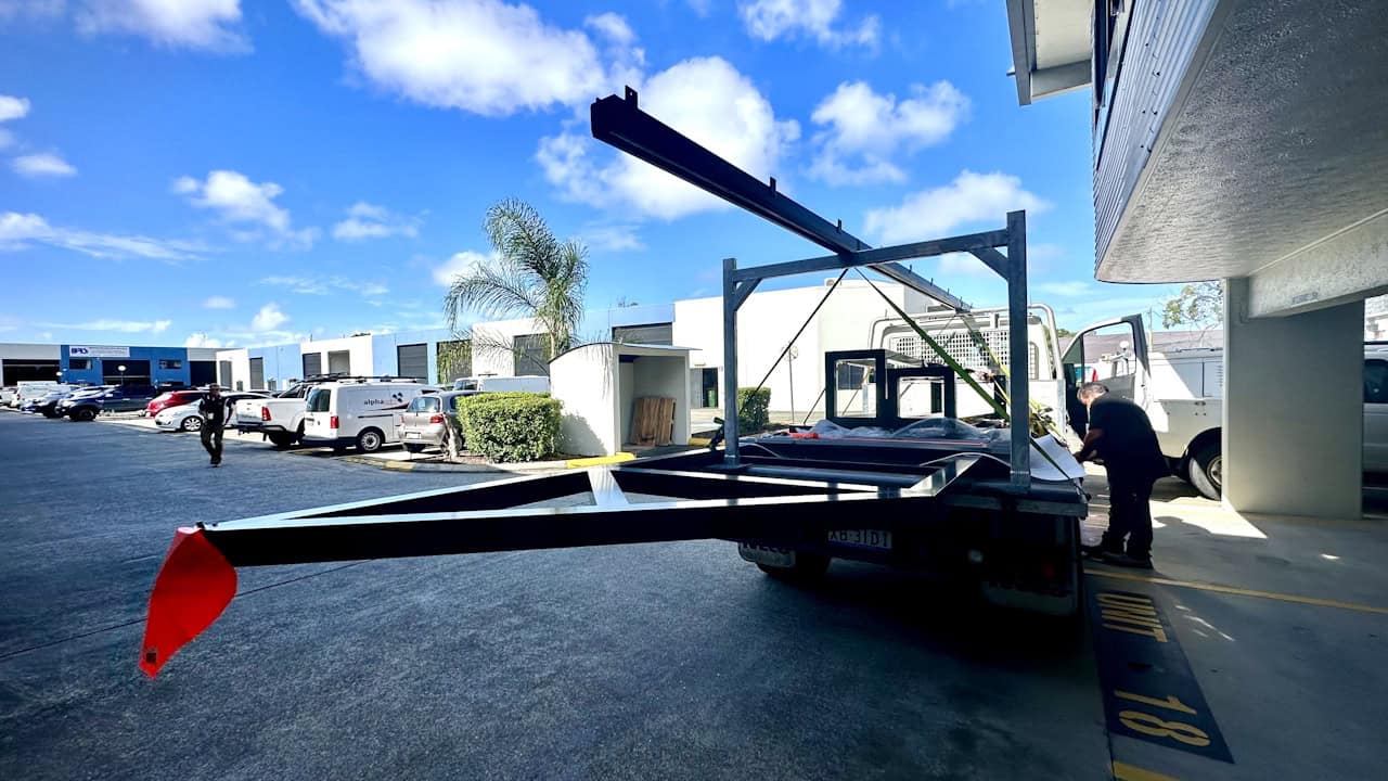 A flatbed trailer with a dark metal frame structure and red flag is parked at a commercial or industrial facility under a blue sky with white clouds.