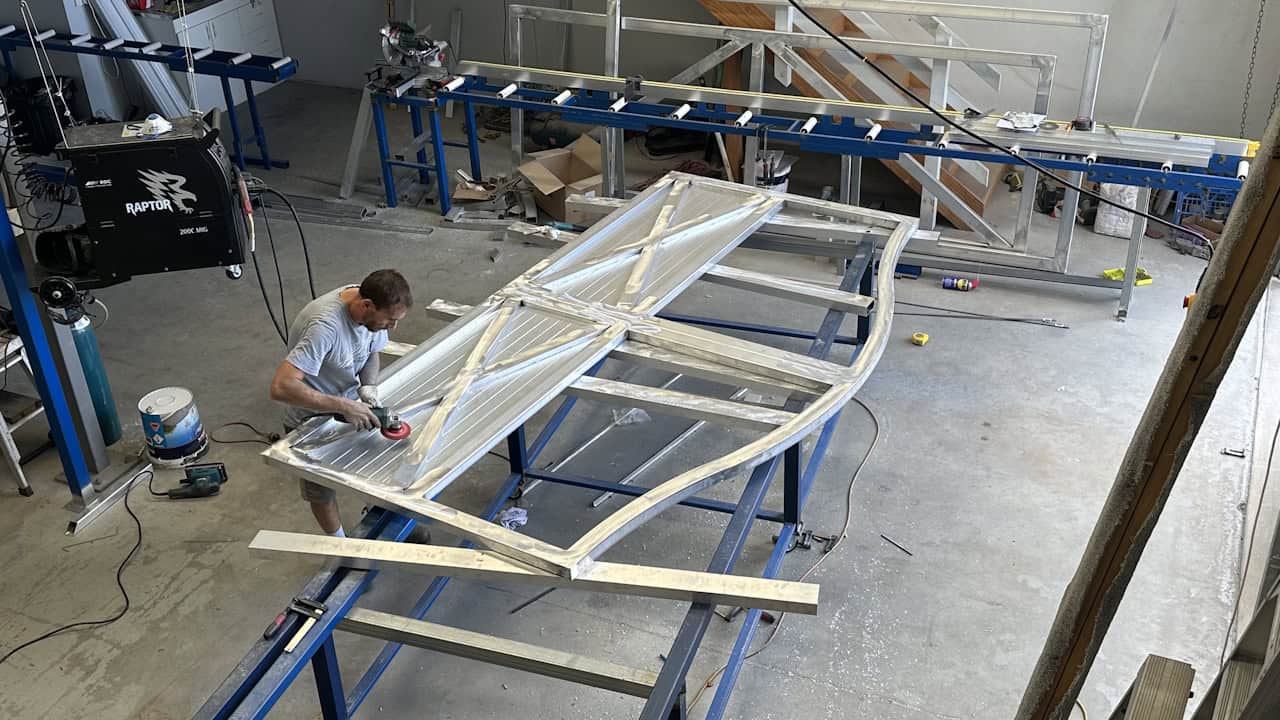 A person works on a large wooden frame structure with curved elements in a workshop, using power tools. Blue work tables and equipment are visible in the industrial space.