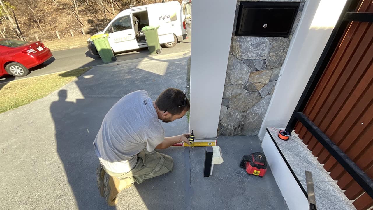 A man kneels on a porch, using a level tool to measure or install something near a white pillar with stone detailing, while a white van with "alpha" text is visible in the street behind him.