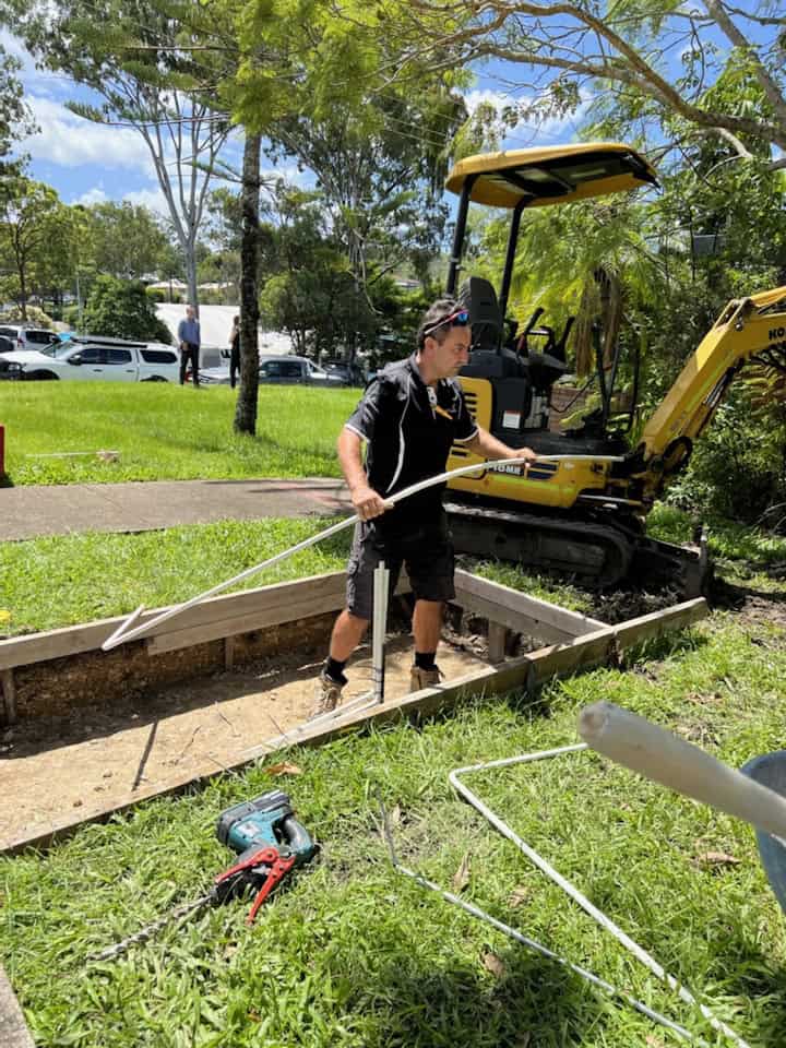 A construction worker in black clothing operates measuring equipment near a wooden frame structure, with a yellow mini excavator positioned behind him on a grass lawn surrounded by trees.