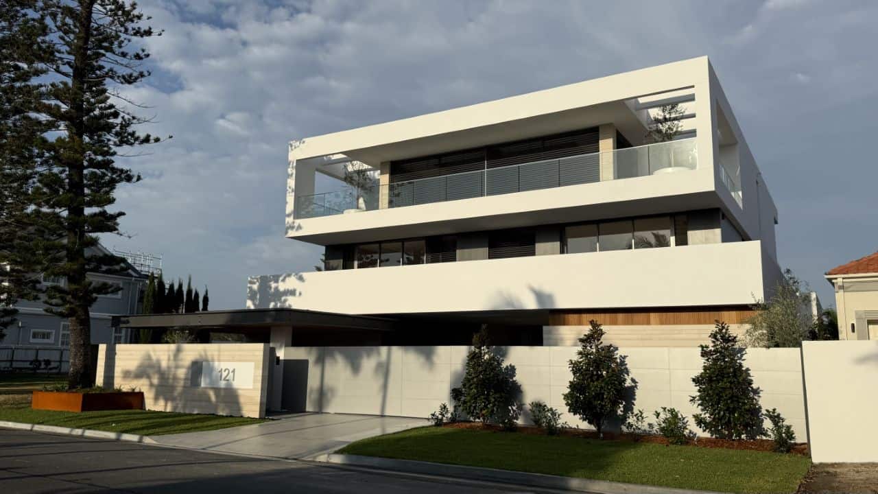 A modern three-story white house with geometric horizontal layers and glass balconies stands behind a manicured lawn with young trees. The number "121" appears on the front wall near the garage.
