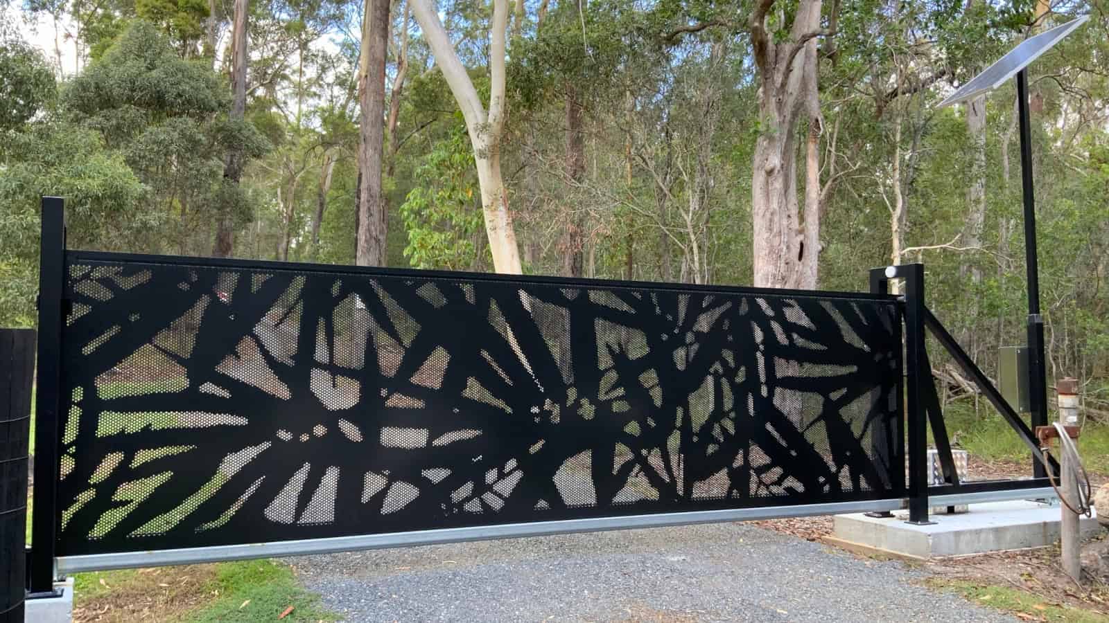 A black decorative solar powered trackless cantilever gate with a complex cut-out pattern, installed at the entrance of a property in Redland Bay, SE QLD, with trees in the background.