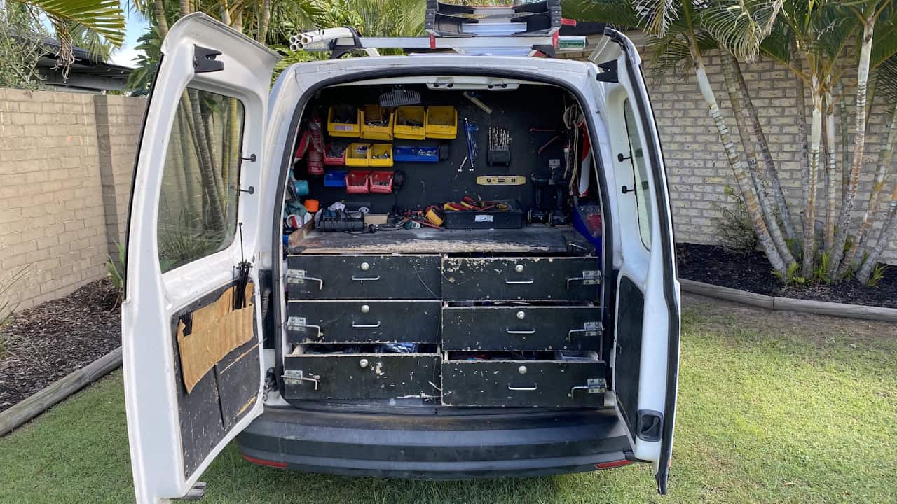 A white work van with open rear doors displays a custom storage system featuring black drawers and colorful tool organizers, parked on grass beside a brick wall with palm trees.
