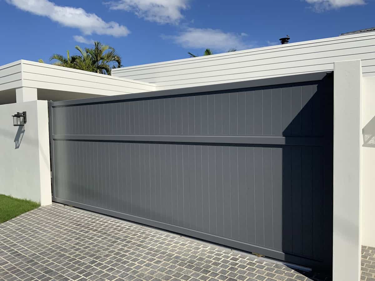 A large modern gray sliding gate spans a driveway entrance, flanked by white walls and a flat roof overhang, with palm trees and blue sky visible behind.