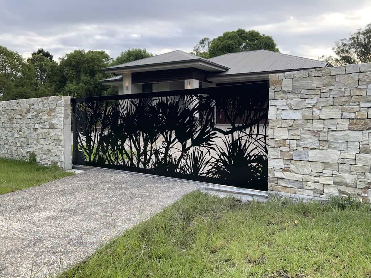 A decorative black metal sliding gate with laser-cut native plant silhouettes spans a driveway entrance between stone walls, with a modern house visible behind under an overcast sky.