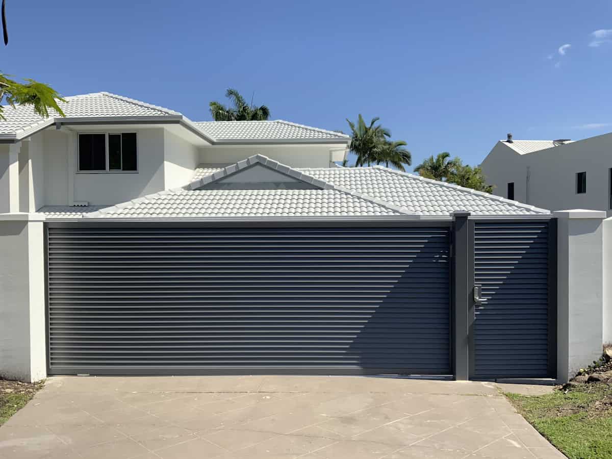 A modern two-story house with white tile roof and cream walls features a dark horizontal-slat garage door and matching pedestrian gate set in a light-colored boundary wall, under clear blue skies with palm trees visible.