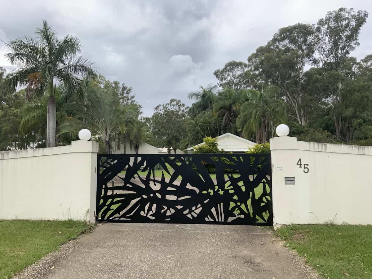A decorative black metal gate with an abstract leaf pattern spans a driveway entrance between cream-colored walls topped with white spherical ornaments. The number "45" appears on the right pillar. Tropical trees and cloudy skies fill the background.