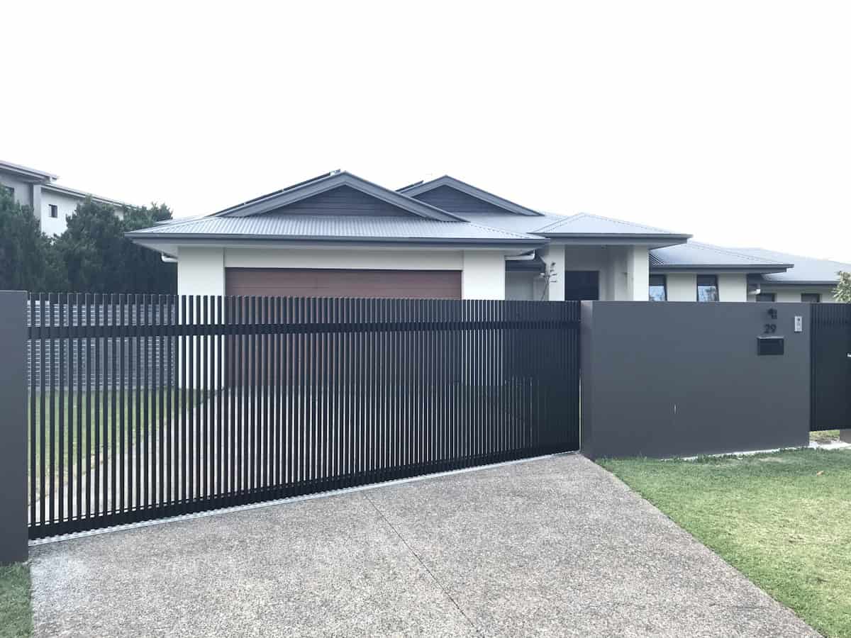 A modern single-story house with white walls and dark roof sits behind a tall black vertical-slat fence and sliding gate, with a concrete driveway leading to the garage.