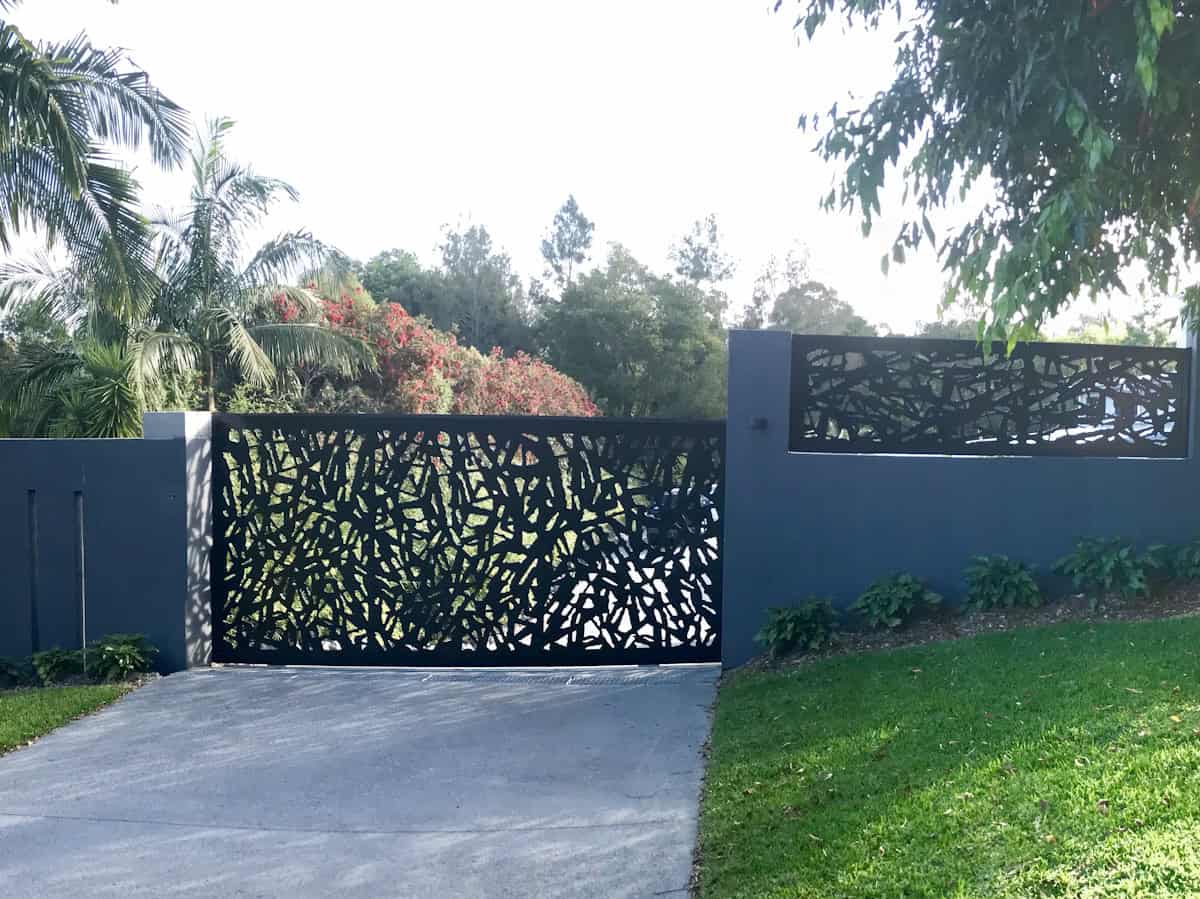A modern black gate with an intricate laser-cut geometric pattern sits between gray walls along a paved driveway, surrounded by green lawn and tropical trees including palms.