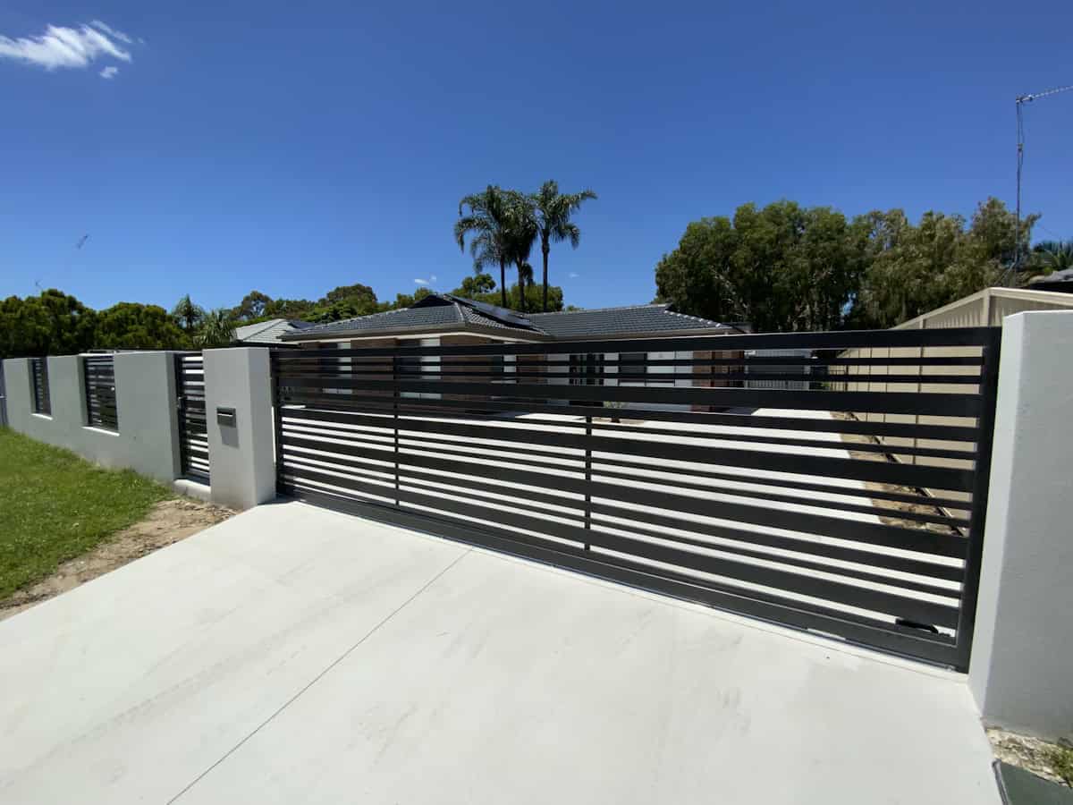 A modern horizontal slat driveway gate in dark metal spans a concrete driveway, flanked by matching fence panels and light-colored pillars, with a residential house and palm trees visible behind.