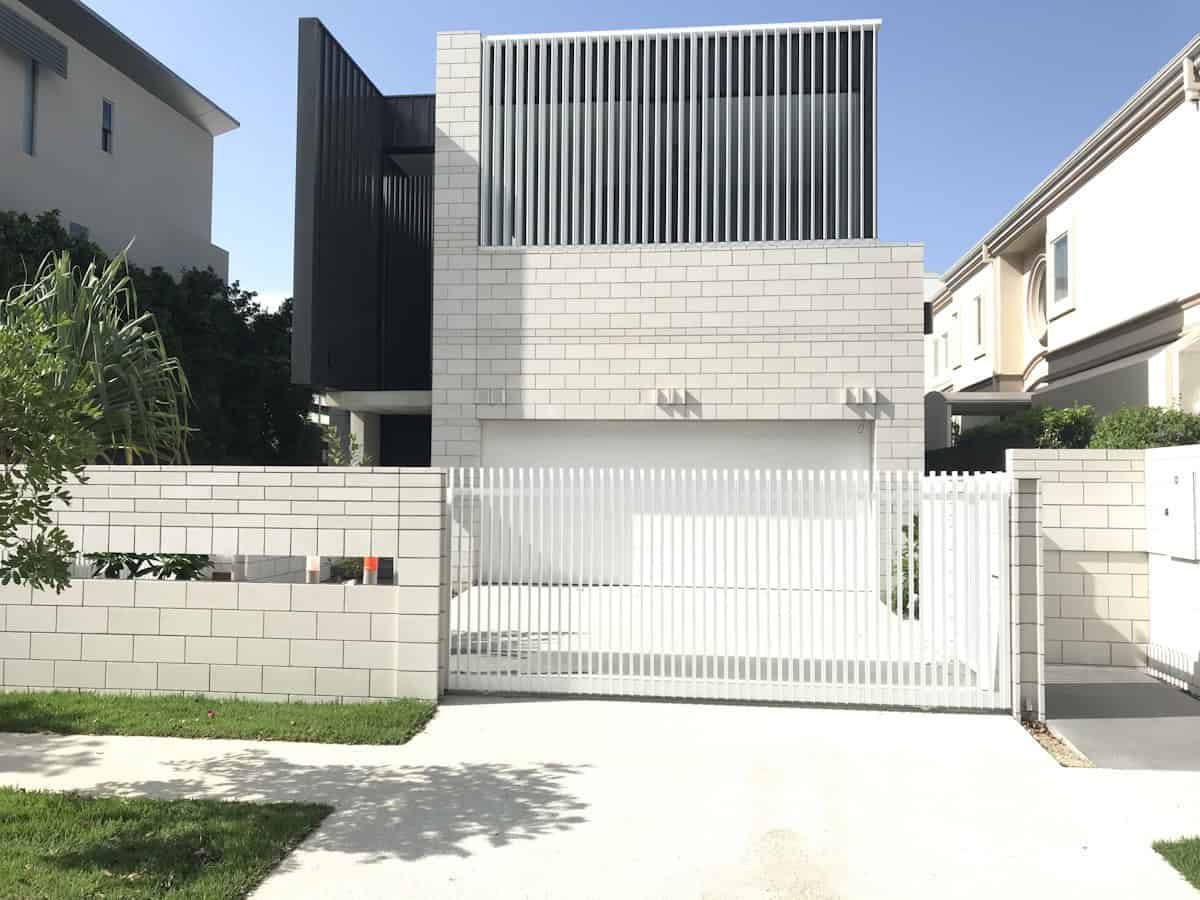 A modern three-story house features vertical white slat screening on its upper level and matching slatted gate, set behind a white brick boundary wall in a suburban street.