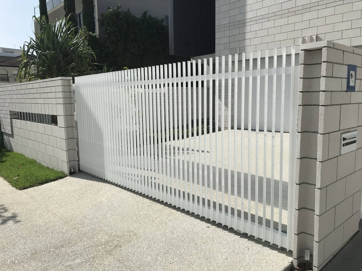 A modern white slatted gate with vertical bars spans between two gray brick pillars, creating a semi-transparent barrier in a contemporary residential setting with manicured lawn and buildings.