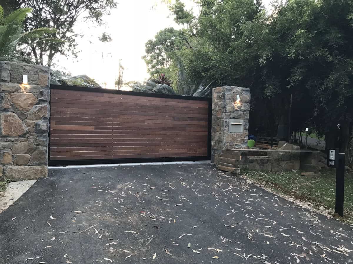 A dark wooden sliding gate spans between stone pillars on an asphalt driveway, surrounded by lush trees and vegetation, with scattered leaves on the ground and an intercom panel visible.