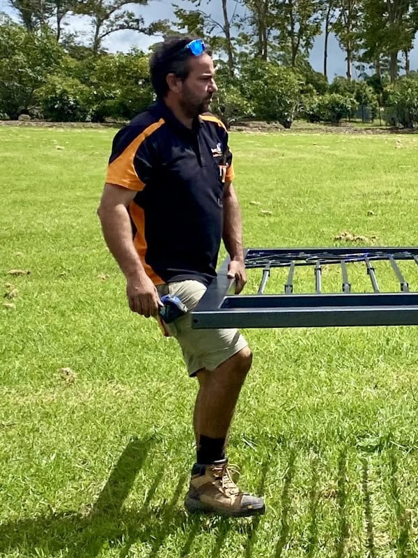 A man in a black and gold polo shirt carries a black metal frame or rack across a grassy lawn, with trees visible in the background on a sunny day.